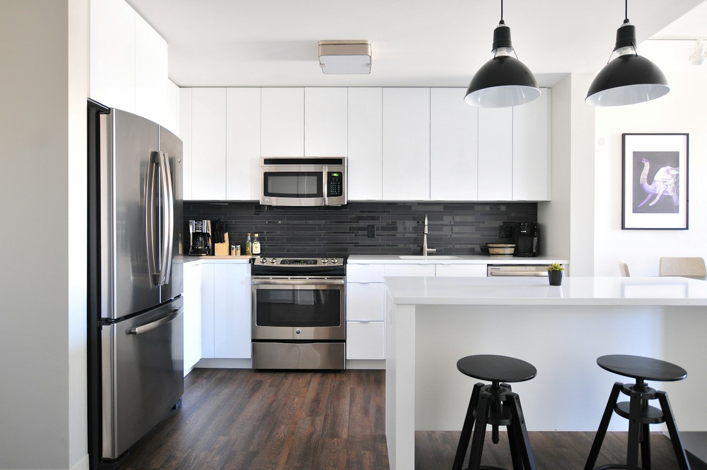 A white kitchen with a steel fridge, a black tiled splashback, and two black stools at the island